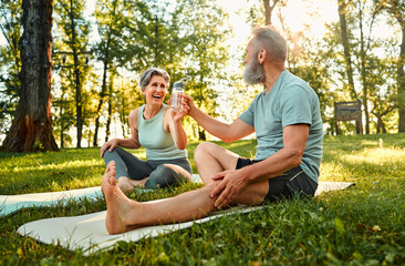 Fototapeta premium Sport and recreation outdoors. Angle view of two family partners having rest and sitting on fitness mats in park. Smiling senior lady sharing bottle of cold fresh water with husband.