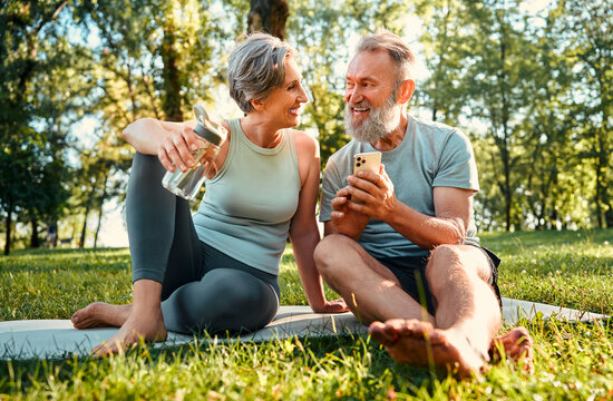 Happy training together. Loving partners resting on mat after physical activities outdoors and looking at each other. Radiant woman holding water bottle while man presenting pictures on cellphone.