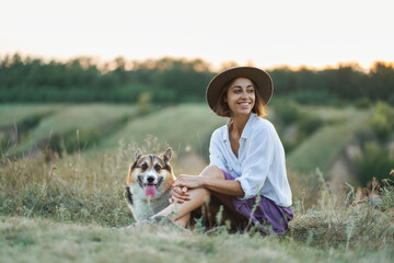 Attractive young stylish woman with her pet welsh corgi dog sits at meadow in nature with grassy hills at sunset