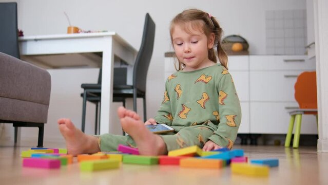 A little beautiful girl sits on the floor in the apartment and plays with her colorful toys. Child at the home enjoing and siling.