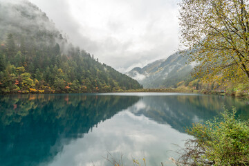 The serene Long Lake mirrors the autumnal beauty in the misty morning at Jiuzhaigou, China, creating a harmonious reflection of tranquil and ethereal elegance.