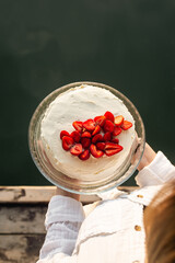 Appetizing white cake decorated with fresh strawberries in female hands on the background of the river, the concept of birthday celebration in nature, top view.