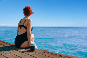 Red-haired woman in a swimsuit is sitting on the edge of a wooden pier with a bottle of refreshing lemonade next to her. Attractive girl enjoys the sea view on vacation. Tourism and travel.