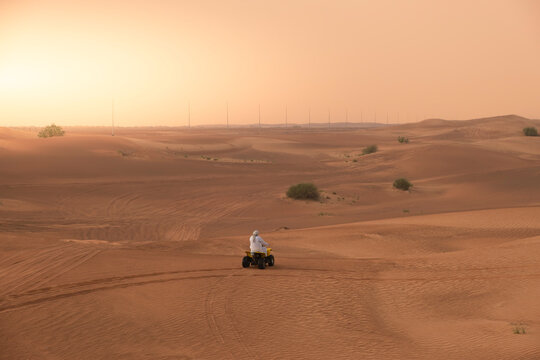 Arab man in traditional white robe outfit and kaffiyeh riding an ATV motorbike alone over sand dunes in the middle of the desert.