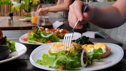 Man eating full meal as poached egg, salad and avocado toast in cafe. Close-up of man with fork pierces poached egg and eat tasty yolk. Order poached egg to eat healthy full meal. Full meal concept.