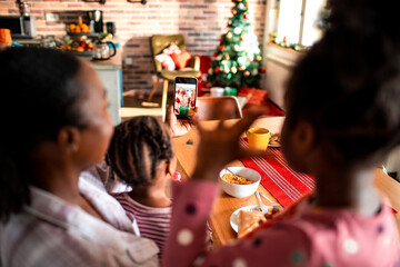 Young African American mother talking to Santa with her children on a video call on a smartphone during the Christmas and new year holidays at home