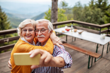 Senior Caucasian couple taking a selfie on smartphone on a balcony of a cabin house in the forest