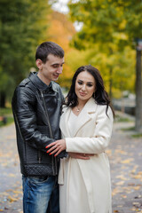 Fototapeta premium A man hugs his pregnant wife. A man in a black leather jacket hugs and kisses a woman in a white coat. Autumn