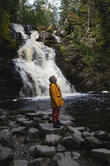 Obraz premium a young beautiful woman tourist stands in a yellow jacket against the backdrop of a waterfall surrounded by forest