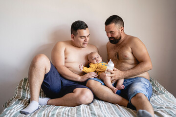 Family of gay couple feeding formula milk to their adopted son in their bedroom bed