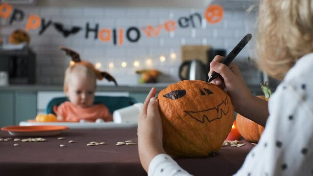 Little girl painting scary face on Halloween pumpkin while her cute baby sister trying on headband with bat wings on the blurred background, selective focus