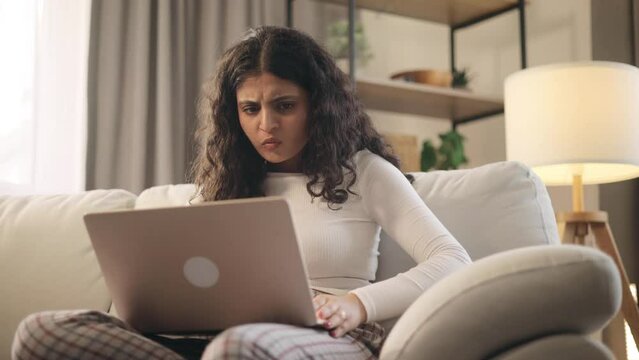 Unhappy young curly woman feeling annoyed when getting message on laptop computer sitting on couch at home Nervous female having problem conflict online indoors. High quality 4k footage