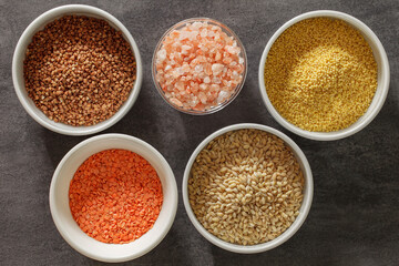Buckwheat, millet, pearl barley, red lentils and pink salt in bowls on dark background, top view