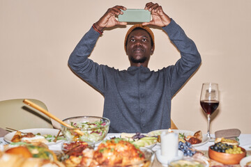 Front view portrait of young African American man taking photo of Thanksgiving dinner table shot...