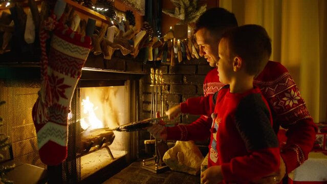 Young father with cute boy using bellows to blow air into fireplace. Dad and son together by cozy fireplace with hanging stockings and Christmas garland lights. Family at Winter and Christmas holidays