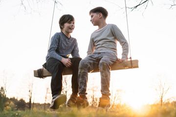 Two wonderful boys sitting on board of swings and discussing impressions of funny leisure outdoors, swinging at sunset.