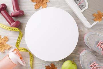 Autumn weight reduction journey. Top view shot of weight scale, shoes, tape measure, dumbbells, green apple, water bottle, dry autumn leaves on light wooden background with blank circle for ad or text