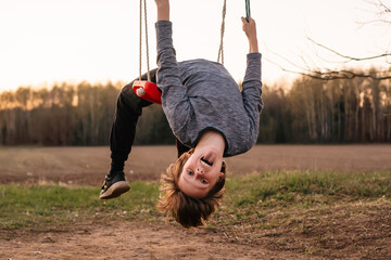 Teen boy sitting on swings, bending back, creating arch and looking at camera with stick tongue...