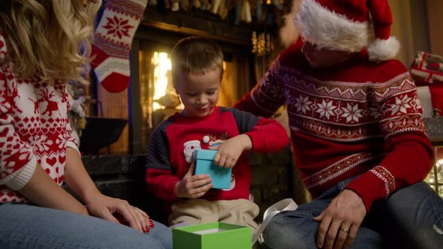Excited cute kid unwrapping gift box. Super happy little boy excited to get surprise present near decorated Christmas tree. Happy family sitting at fireplace. Cinematic Christmas eve RED camera