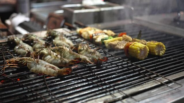 Peeled shrimps and vegetables, corn are cooked on an open flame. A man flips shrimp on the grill, seafood is on fire.