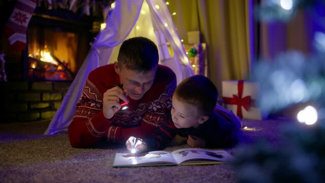 Cinematic RED camera shot father and boy playing with flashlights in living room in canvas tent with Christmas tree and stockings decoration on fireplace burning on blurred background, slow motion 4K