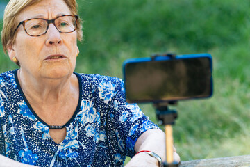 Senior woman recording a podcast and vlogging with smartphone sitting on a park bench