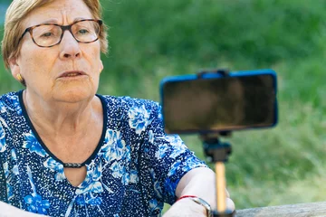Fototapeten Senior woman recording a podcast and vlogging with smartphone sitting on a park bench © MiguelAngel