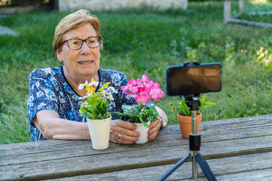 Senior woman recording a podcast and vlogging with smartphone, sitting on a park bench, with flowers on the table.