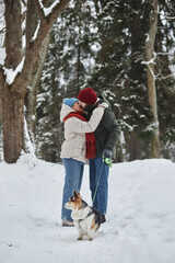 Fototapeta na wymiar Vertical full length portrait of romantic young couple kissing outdoors in winter forest and walking cute dog