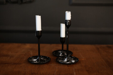 Modern three black glass candlesticks with white candles on wooden table in antique style on dark wall background. Minimalist elegant decor with candle holders with wax different size.
