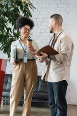 cheerful african american woman holding coffee to go and chatting with asian coworker during break