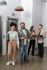 Fototapeta premium happy african american woman and indian man looking at camera near office elevators, diverse people
