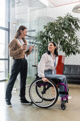 inclusion concept, happy woman holding coffee and talking with disabled colleague in wheelchair