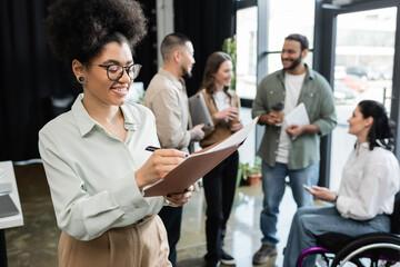 cheerful african american woman taking notes and standing near interracial team in coworking