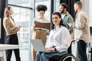 happy disabled businesswoman in wheelchair using laptop near blurred multiethnic colleagues - Powered by Adobe