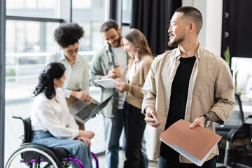cheerful asian man holding smartphone and folder near multicultural team on blurred background