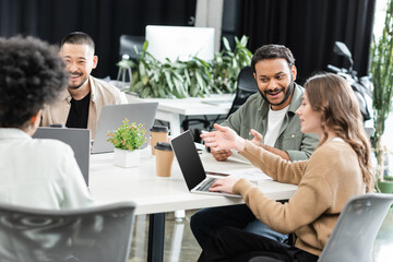 cheerful interracial colleagues using laptops and planning project together in modern coworking