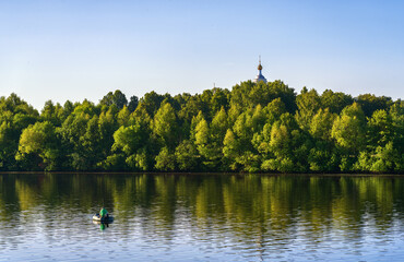 A fisherman on an inflatable rubber boat is fishing opposite an island overgrown with trees over...