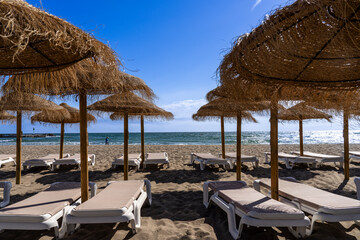 A sunny horizontal shot from under a row of thatched straw parasols on a sandy beach, looking out at empty white sun loungers, the blue sea, and a clear sky. © Hans Hansen