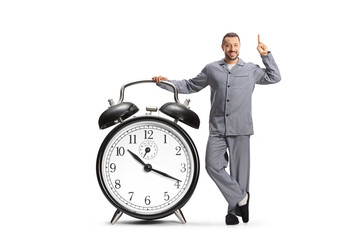 Young man in pajamas leaning on a big alarm clock and pointing up