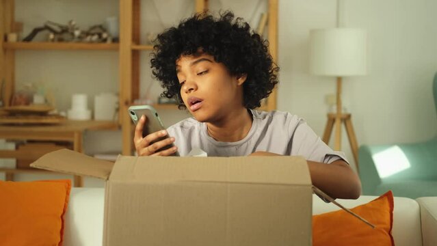 Unhappy angry african woman unpack delivered box receiving damaged shipping mistake problem in postal service. Annoyed confused customer client receiving wrong parcel checking online app on smartphone