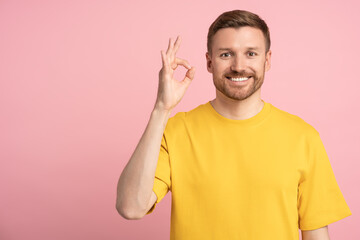 Positive happy man showing OK sign hand gesture on pink background with copy space looking at...