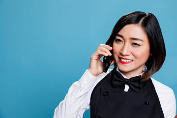 Hotel smiling cheerful asian woman receptionist wearing uniform speaking on smartphone. Joyful waitress answering call and chatting with catering service customer on mobile phone