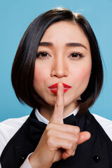 Naklejka premium Serious asian woman in waitress uniform holding forefinger on lips while showing tsss gesture and looking at camera. Young receptionist asking for silence closeup portrait