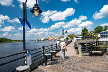 woman in a hat walks along the sidewalk along the waterfront of the Cape Fear River, Wilmington, NC