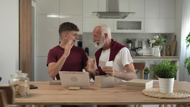 senior man grandfather pensioner gray hair and beard with his grandson teenager boy receive presents in box open read card happy smile in front of digital tablet at home online video call slow motion