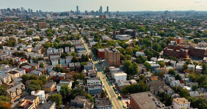 Descending above the sunny scenery of Cambridge, USA. Panorama of the city with usual low-rise buildings with trees among them. Skyscrapers at backdrop.