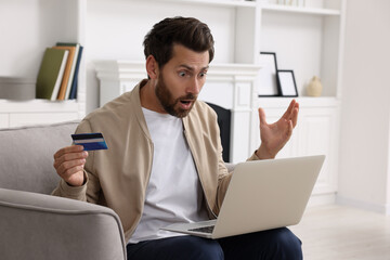 Shocked man with laptop and credit card in armchair at home. Be careful - fraud