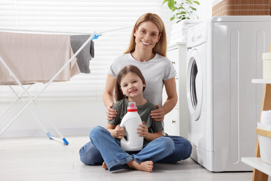 Mother and daughter sitting on floor near washing machine and holding fabric softener in bathroom