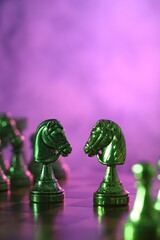 Two knights and other chess pieces on checkerboard in color light, selective focus. Space for text